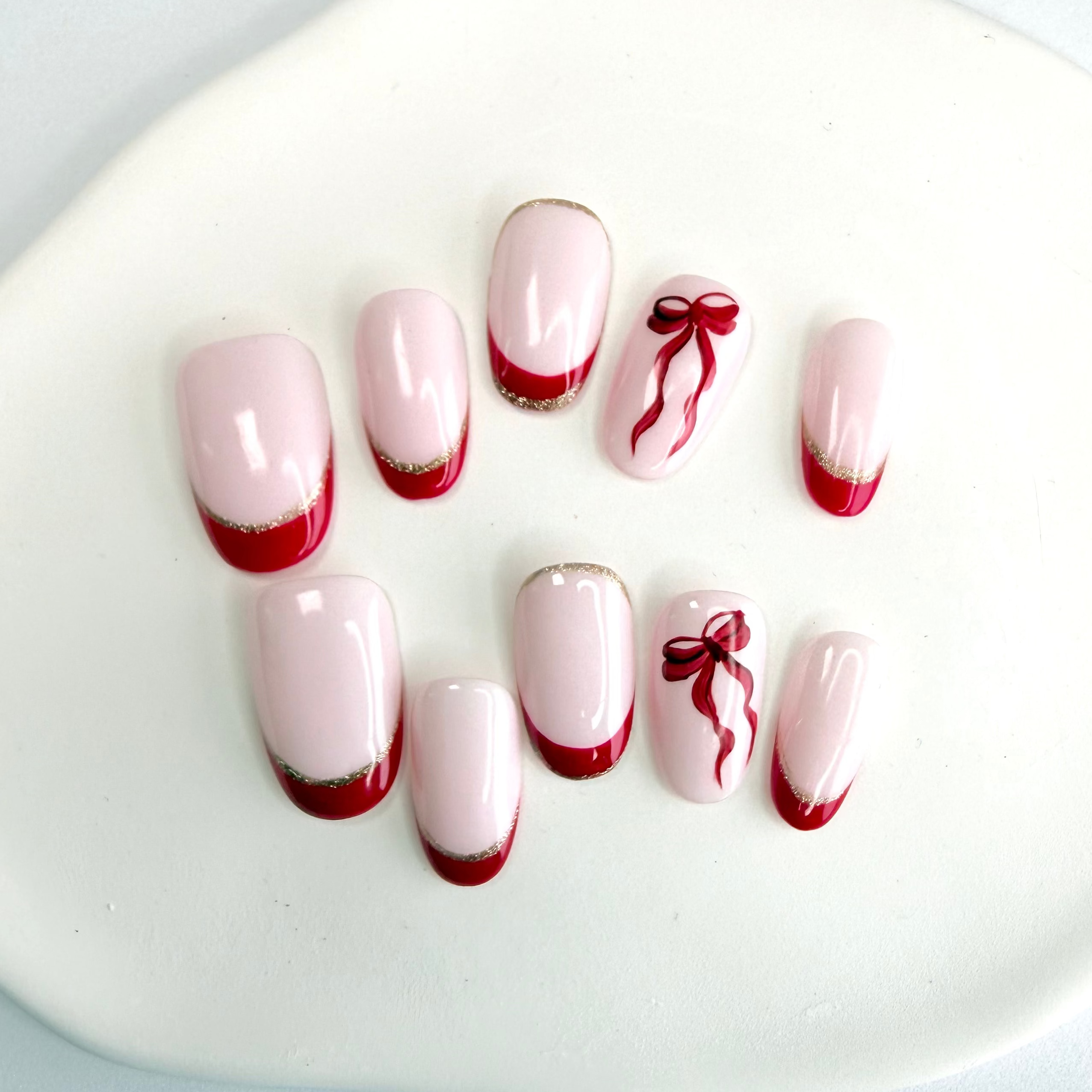 Handcrafted nude press-on nails with red and gold French tips and a red ribbon accent, designed by Vlei Nails Cape Town.