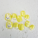Yellow and white kiddies press-on nails with bunny face and heart designs on a textured white background.
