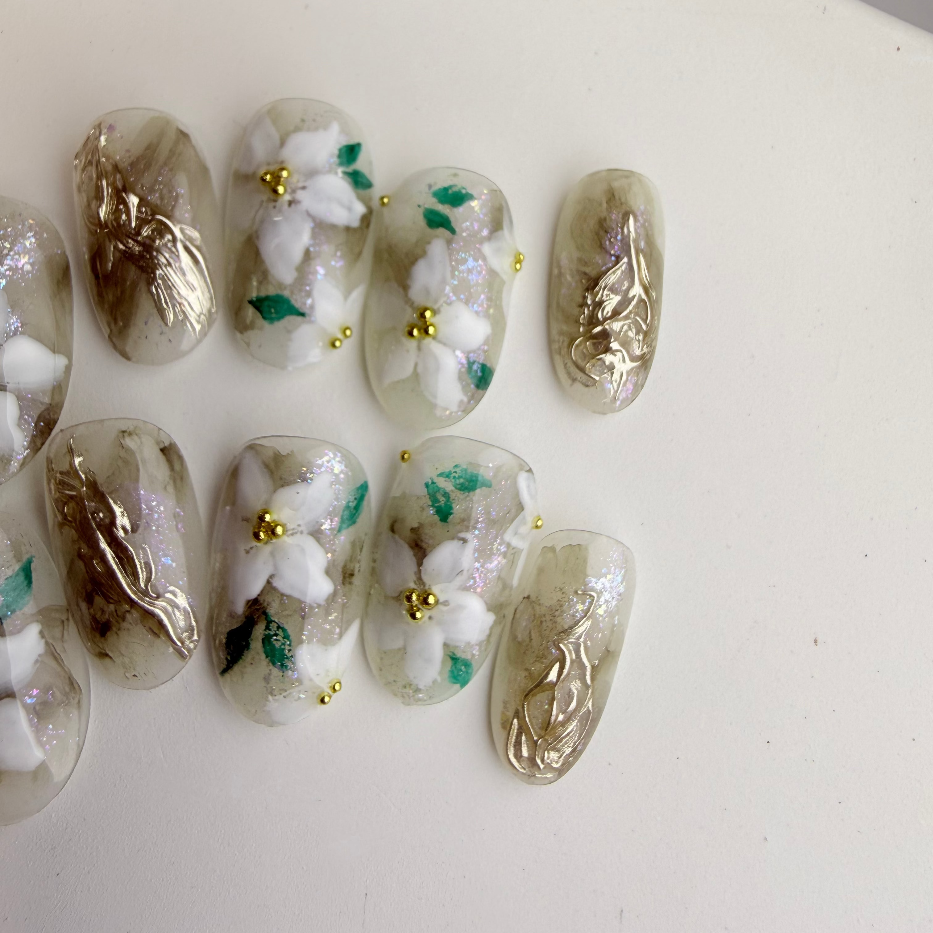Handcrafted green marble press-on nails with white 3D floral designs and gold foil accents.