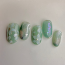 Mint-green press-on nails with white floral designs, marble shimmer and iridescent glow accents.
