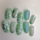 Mint-green press-on nails with white floral designs, marble shimmer and iridescent glow accents.
