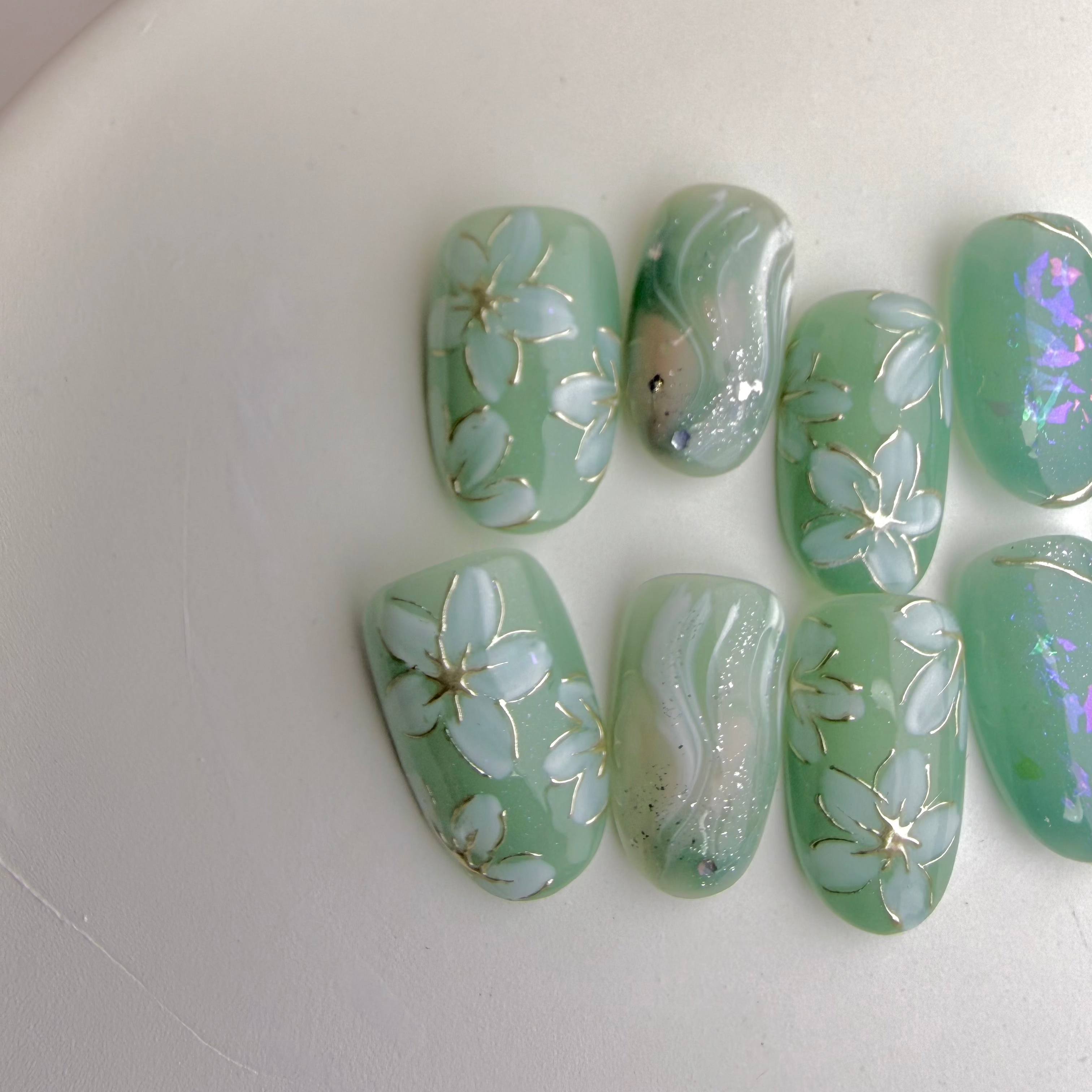 Mint-green press-on nails with white floral designs, marble shimmer and iridescent glow accents.
