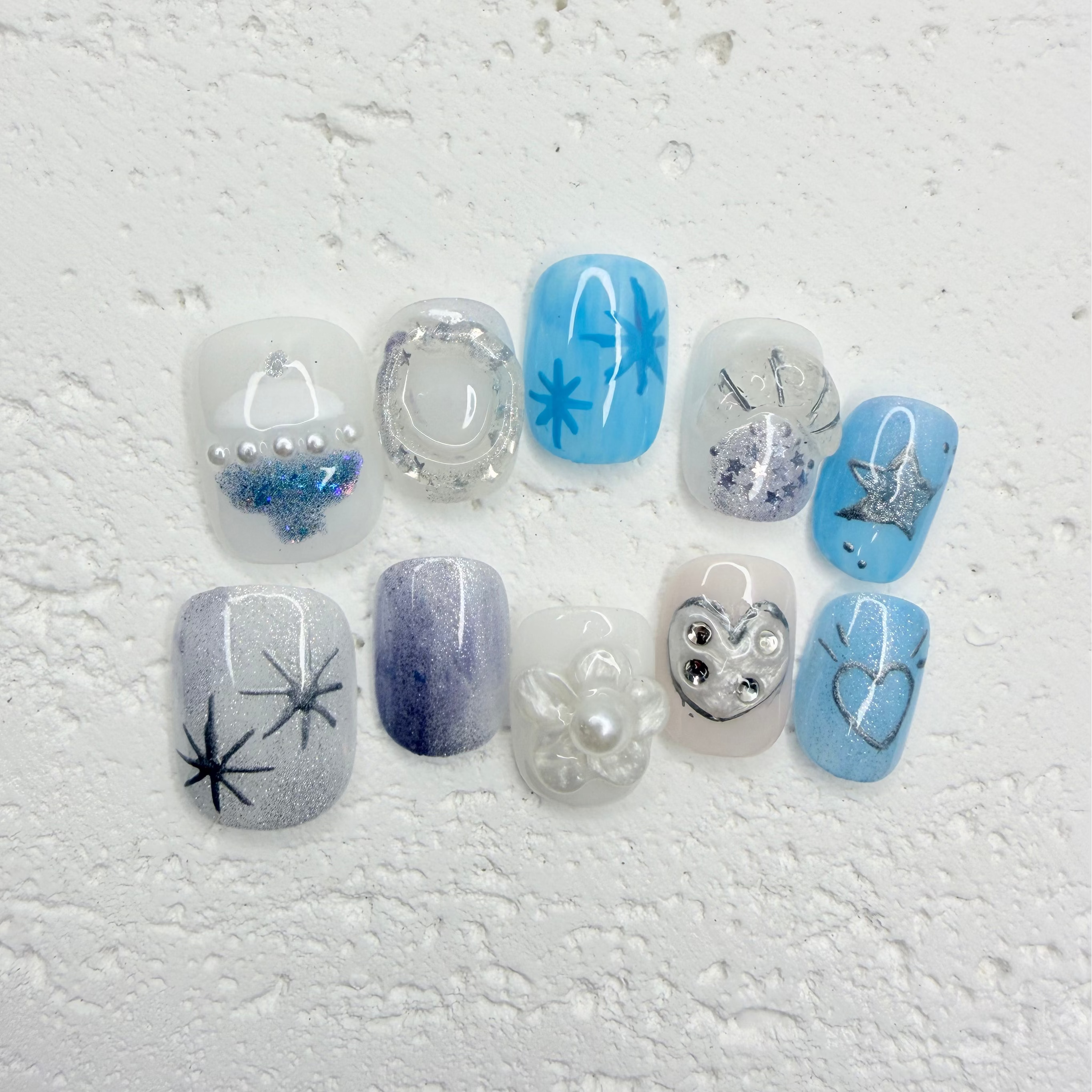 Icy blue and silver press-on nail set with starburst designs, glitter accents, pearls, rhinestones, and a heart outline nail.