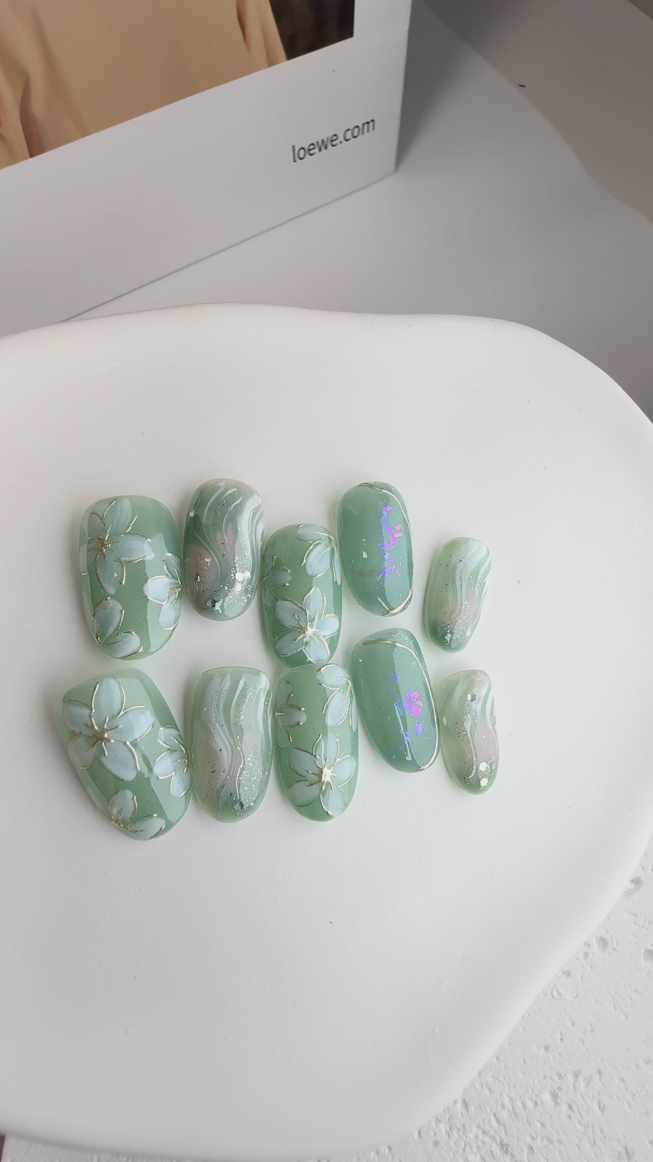 Mint-green press-on nails with white floral designs, marble shimmer and iridescent glow accents.
